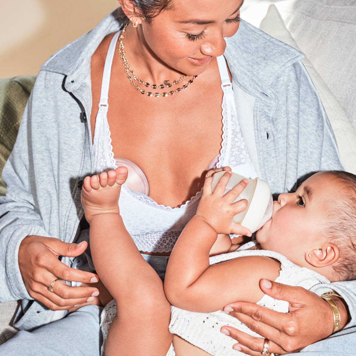 Woman holding a baby that's drinking from a bottle while wearing a Muu pump in her shirt.